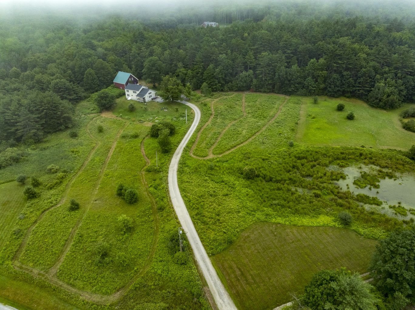 Aerial view of a farmhouse at the end of a winding drive across a fog covered field.
